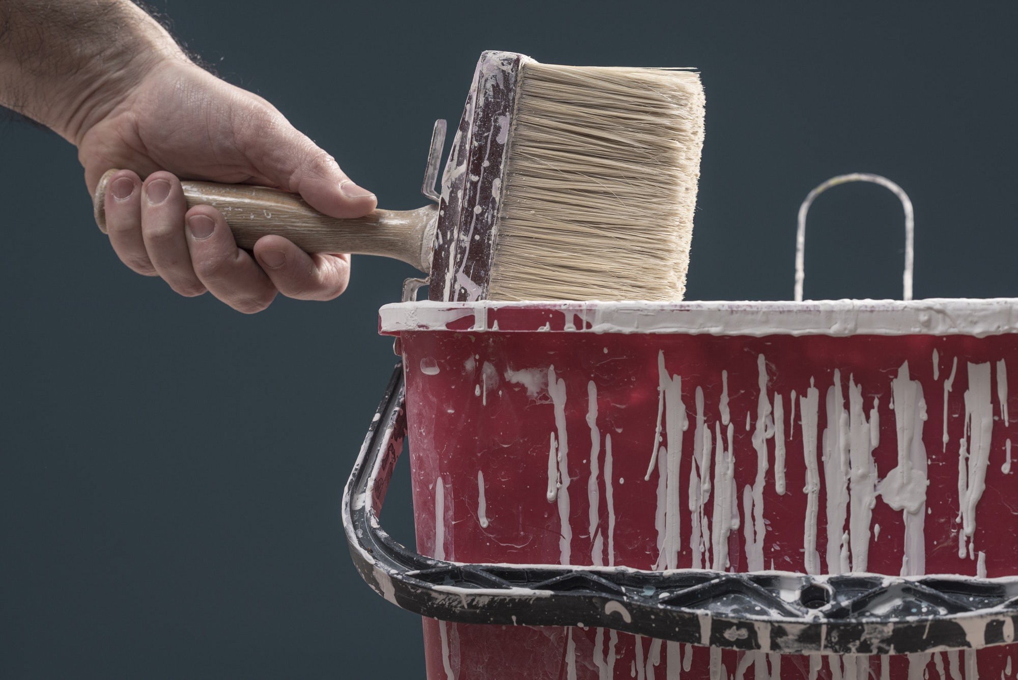 Hand Holding Paint Brush Over Red Bucket of White Paint